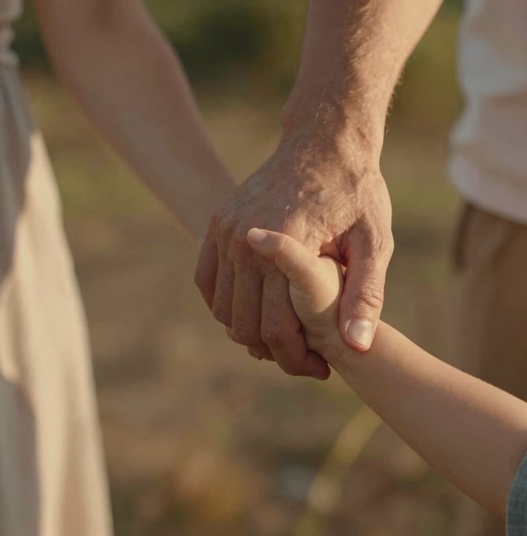 A close-up, cinematic shot of a mother and father's hands holding their toddler's small hand. The skin tones are warm and glowing under a late afternoon sun. The background is a soft-focus blur of Warm Brown garden elements.