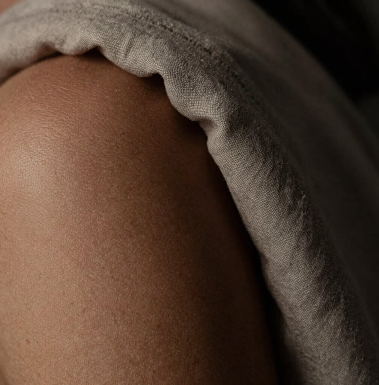 Macro photography showing the delicate texture of skin and a piece of muted sand-colored fabric. The lighting is low-key, creating deep dark charcoal shadows and a sense of calm artistry.