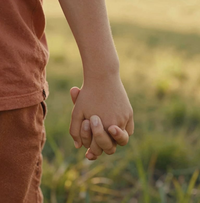 A close-up, artistic shot of a child's hand holding a parent's hand against a background of sun-dappled grass. The lighting is warm and golden. Soft focus, cinematic storytelling style, highlighting textures of skin and terracotta-colored fabric.