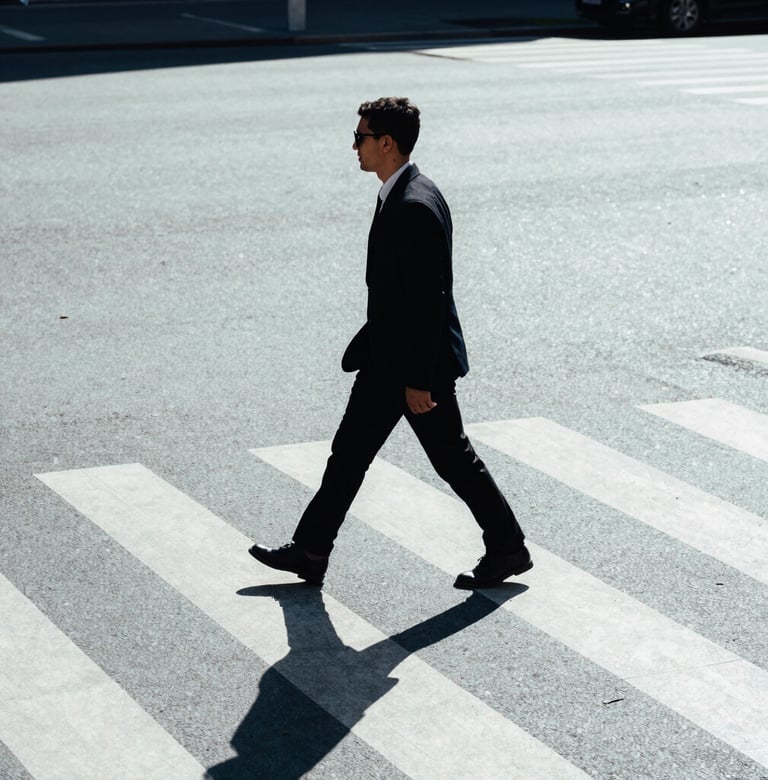 A candid street photograph of a person walking across a zebra crossing in a city. High contrast lighting with long shadows on light gray-blue pavement. Minimalist style, International / Western urban environment.