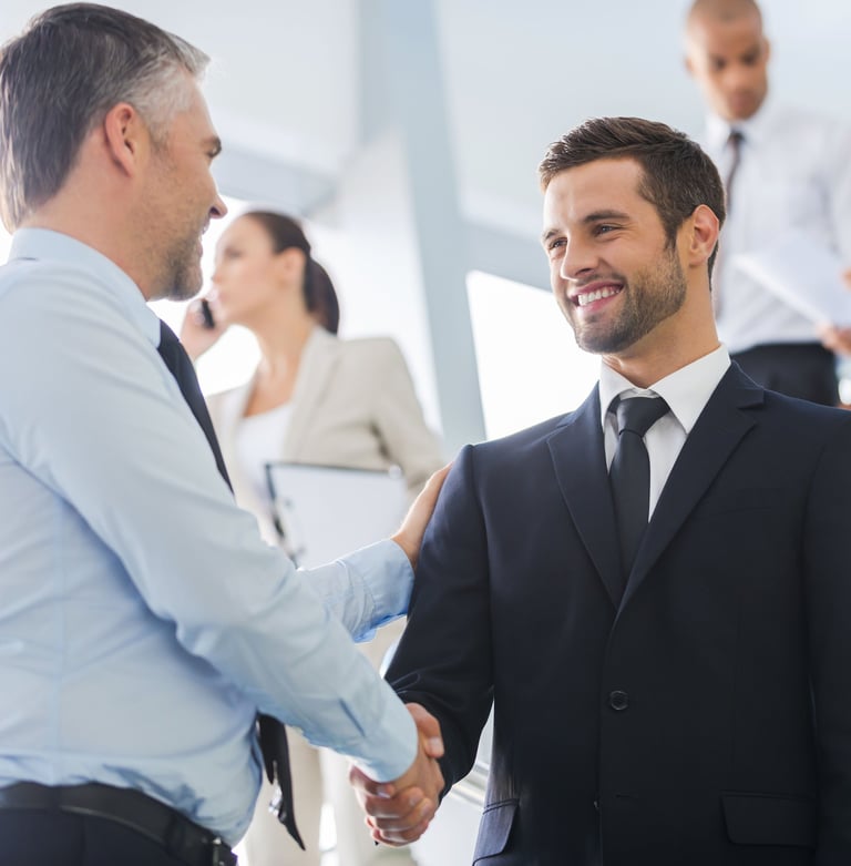Two smiling businessmen in suits shaking hands in a professional office setting.