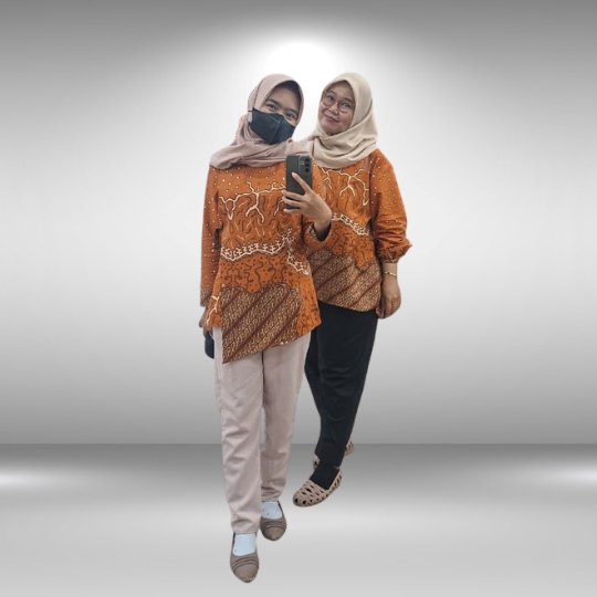 Two women wearing matching orange batik shirts and hijabs posing for a mirror selfie.