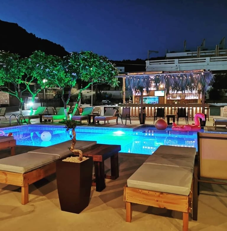 Happy Friends swimming pool at night with a tiki bar, lounge chairs, and illuminated palm trees.
