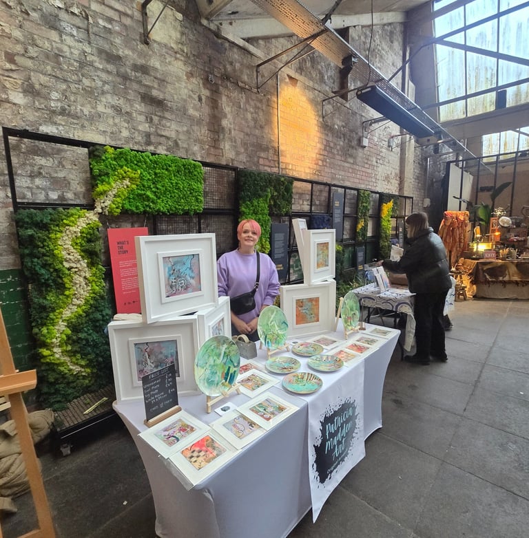 Patricia Madden Artist displaying colorful framed prints and ceramic plates at an indoor art market.