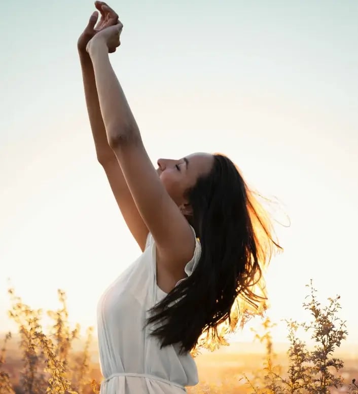 Une femme en robe blanche dans un champ lève les bras vers le ciel, exprimant légèreté et liberté.