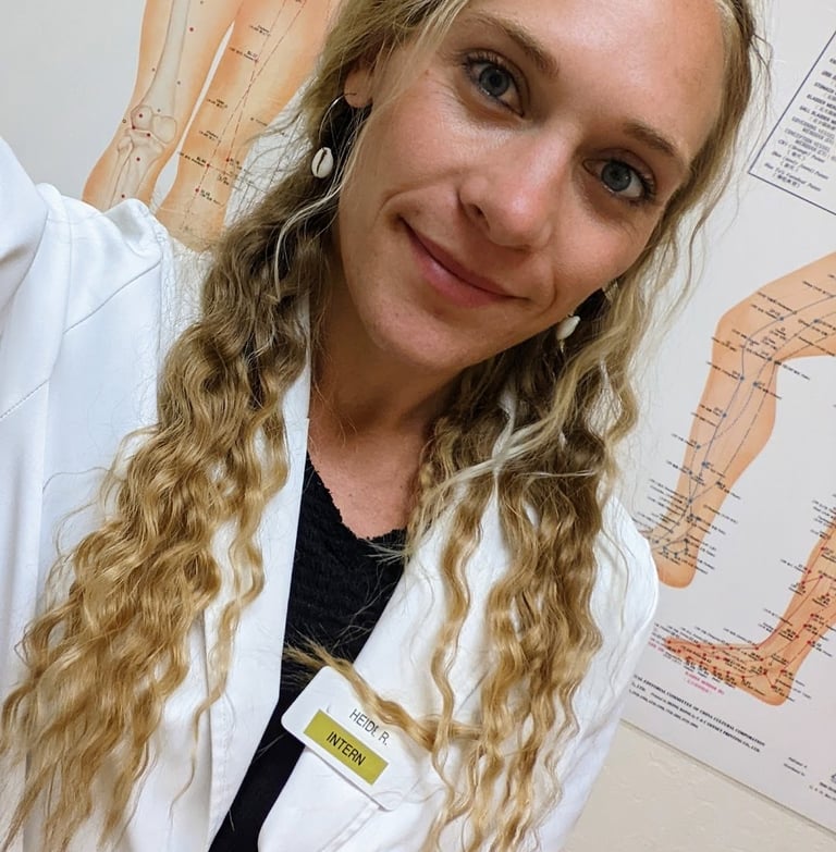 blonde acupuncture intern in a white lab coat posing in front of anatomical meridian charts.