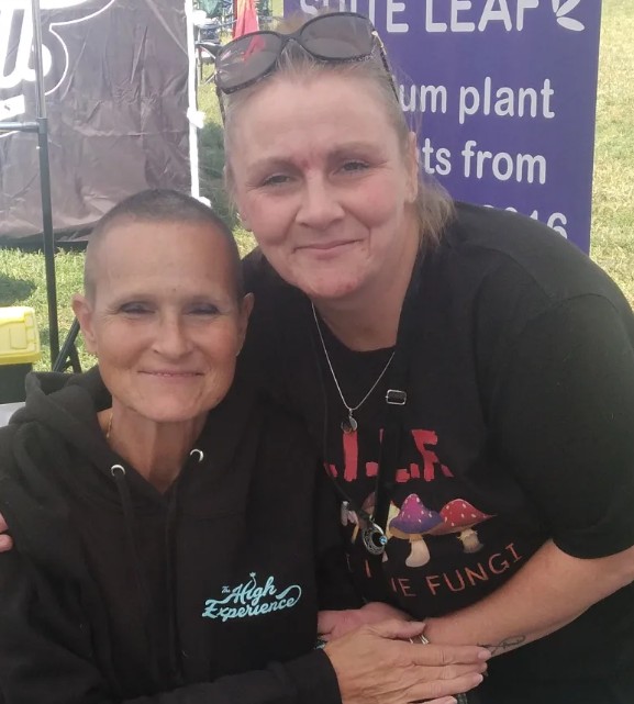 Two women smiling at an outdoor event near a sign for White Leaf plant nutrients.