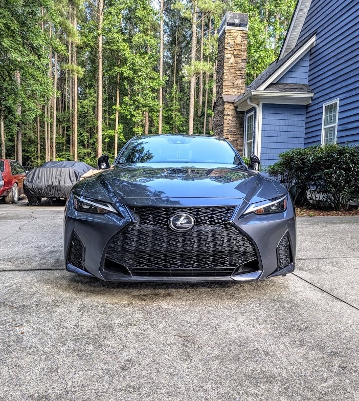 Front view of a charcoal gray Lexus luxury sedan parked on a concrete driveway in front of a blue house.