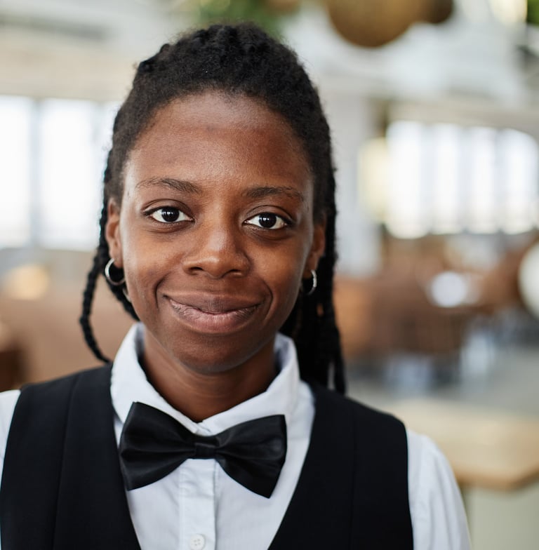 Smiling Black female server in formal uniform with a black bow tie at a modern restaurant.