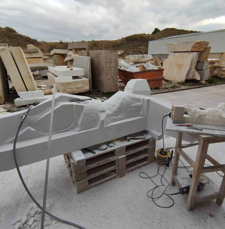 A stone sculpture in progress on a pallet in a masonry workshop with marble blocks and tools.