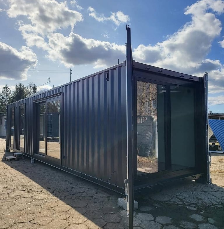 Modern dark grey container home with large glass sliding doors under a bright blue sky.