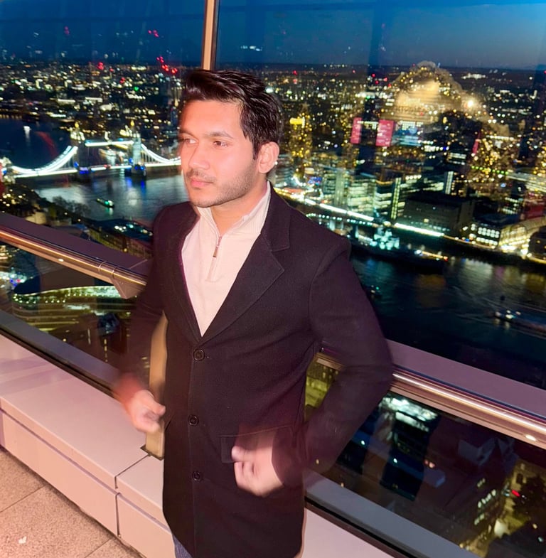 A man in a black coat standing on a London rooftop balcony at night with Tower Bridge in the background.