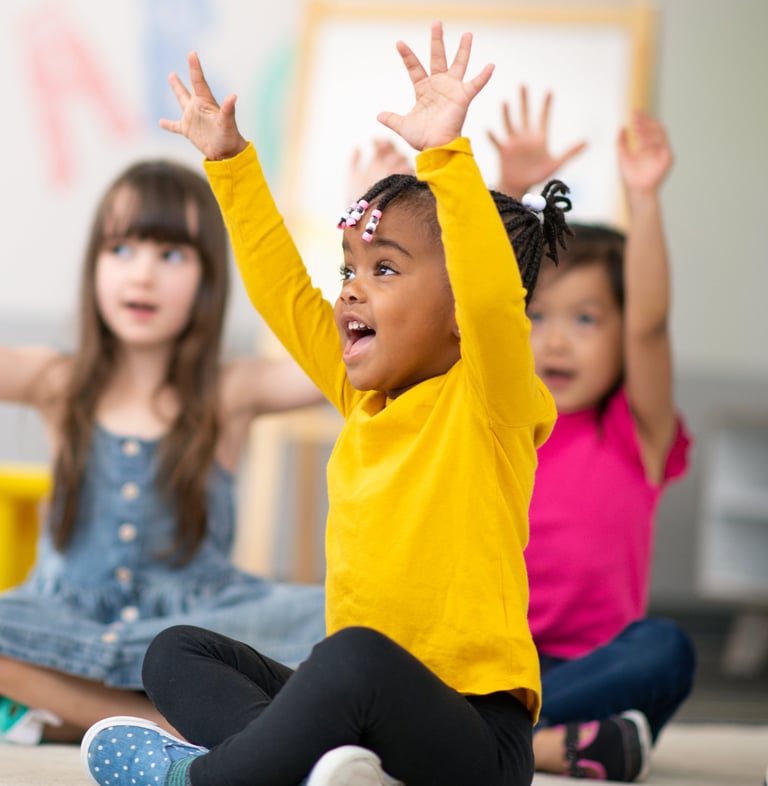 Photo of children in classroom with arms in the air.