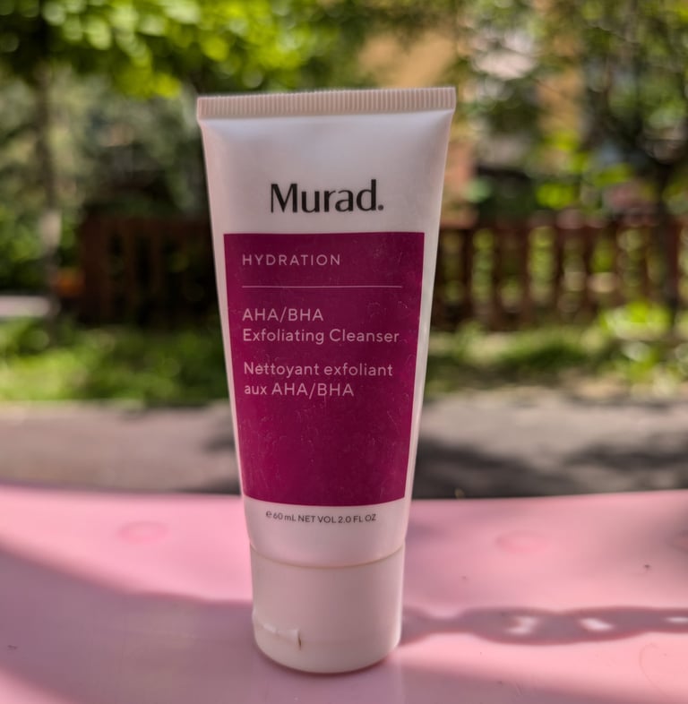 Murad Hydration AHA BHA Exfoliating Cleanser tube on a pink surface with a sunny outdoor background.