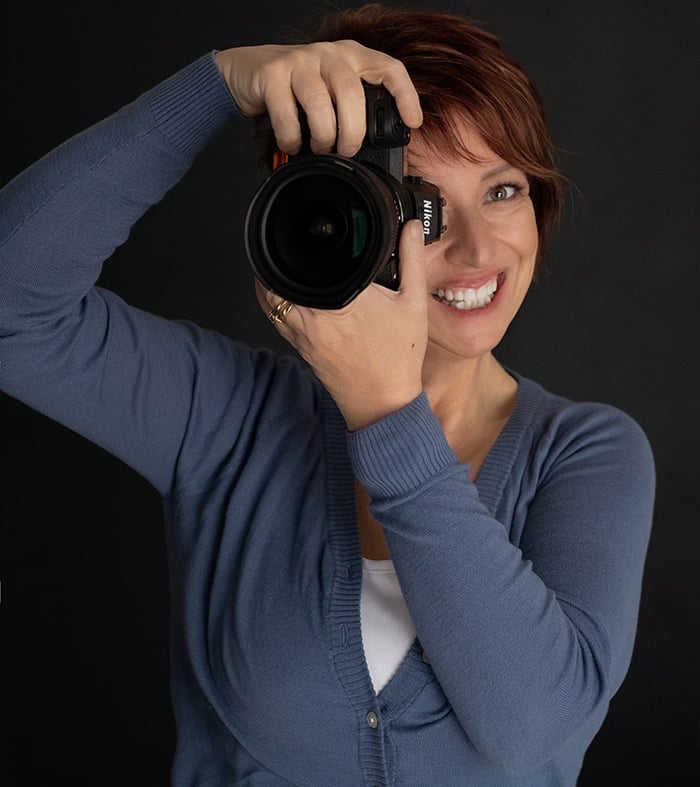 Smiling female professional photographer with red hair holding a Nikon DSLR camera against a black background.