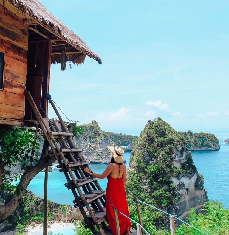 A woman in a red dress climbs a wooden ladder to a Bali treehouse overlooking Nusa Penida islands.
