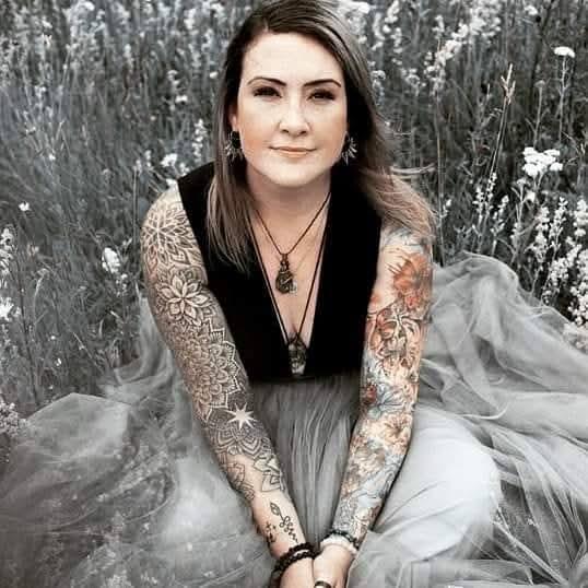 A woman with intricate sleeve tattoos sits in a flower field wearing a tulle dress and bohemian jewelry.
