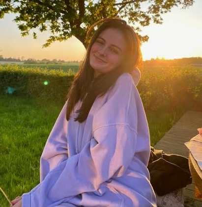 Smiling woman in a purple hoodie sitting outdoors during a golden hour sunset in a green field.