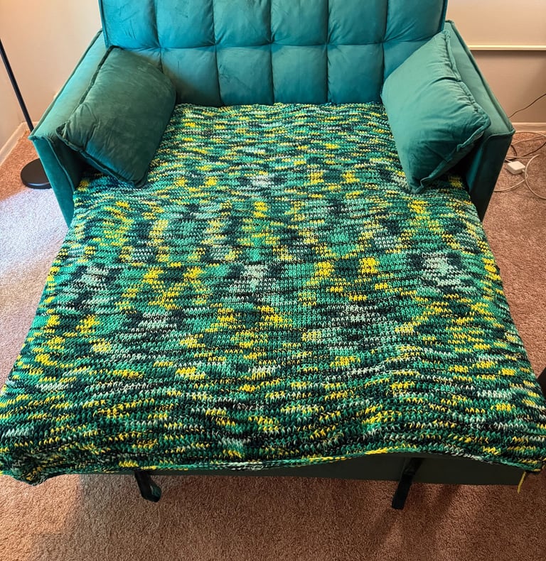 A green velvet convertible sofa bed covered with a handmade green and yellow variegated crochet blanket.