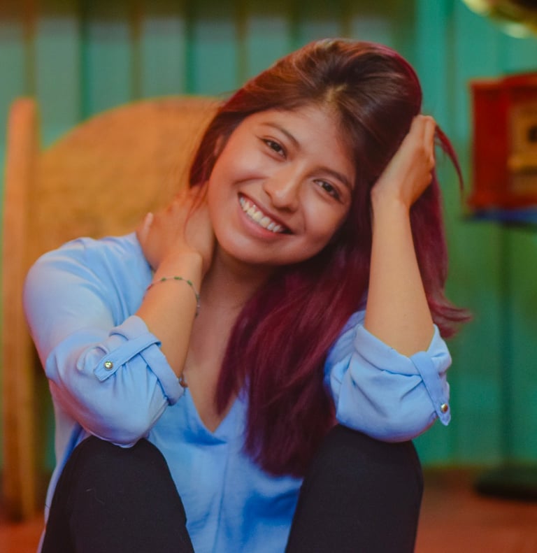 A smiling young woman with dyed hair wearing a light blue shirt in a cozy room.