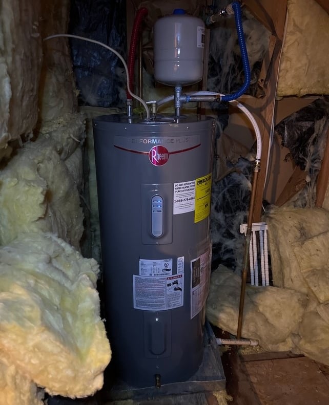 A Rheem Performance Plus electric water heater installed in an attic surrounded by fiberglass insulation.