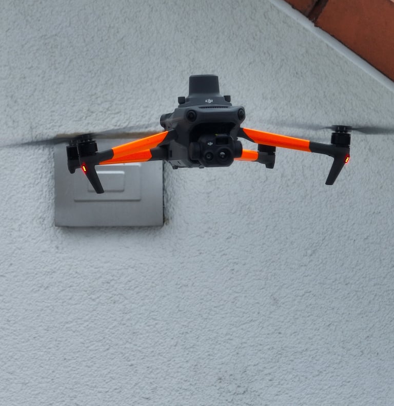 A DJI Mavic 3 Enterprise drone with orange skins hovering near a white building wall.