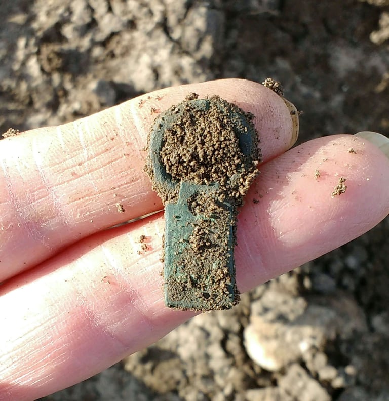 A copper alloy Medieval buckle with green patina, partially covered in soil, held on two fingers.