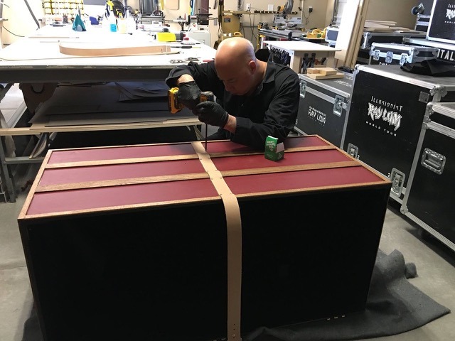 Ray Lum uses a power drill to assemble a custom wooden magic illusion trunk in a workshop.