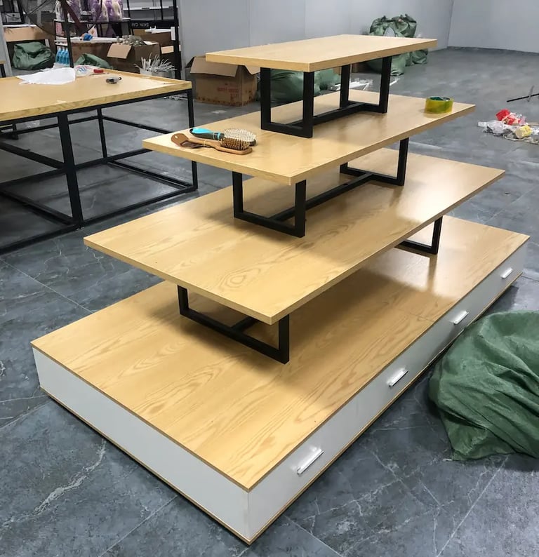 Four-tier wooden retail display table with black metal frames and white storage drawers.