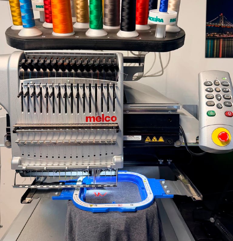 A multi-needle Melco commercial embroidery machine stitching a red logo onto a gray garment.