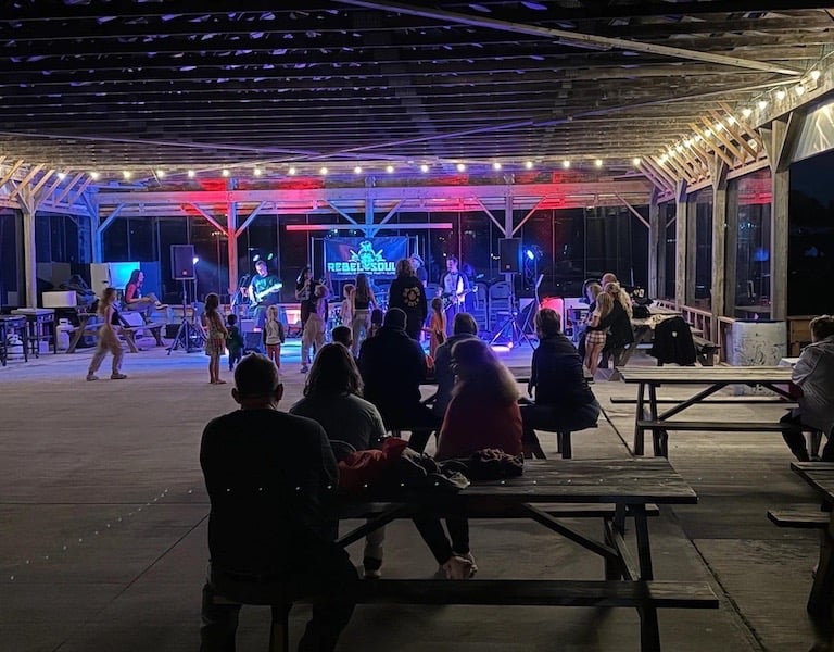Live band playing for an audience under a lit pavilion with picnic tables at an outdoor concert event.