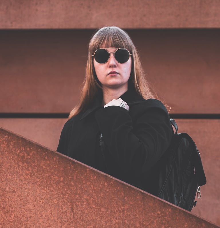 a woman in sunglasses and a black jacket standing in between a wall and a staircase