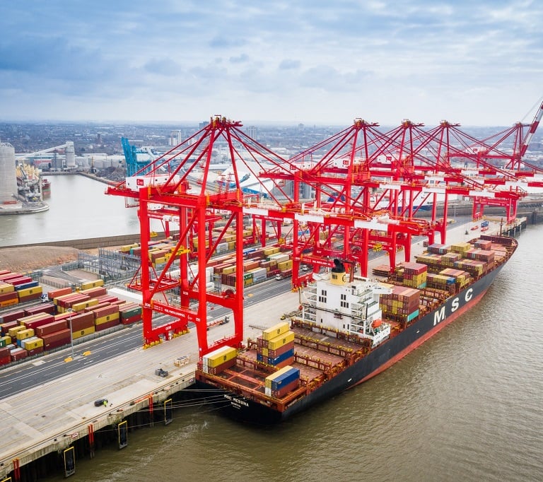 An aerial view of an MSC cargo ship docked at a port with large red gantry cranes loading shipping containers.
