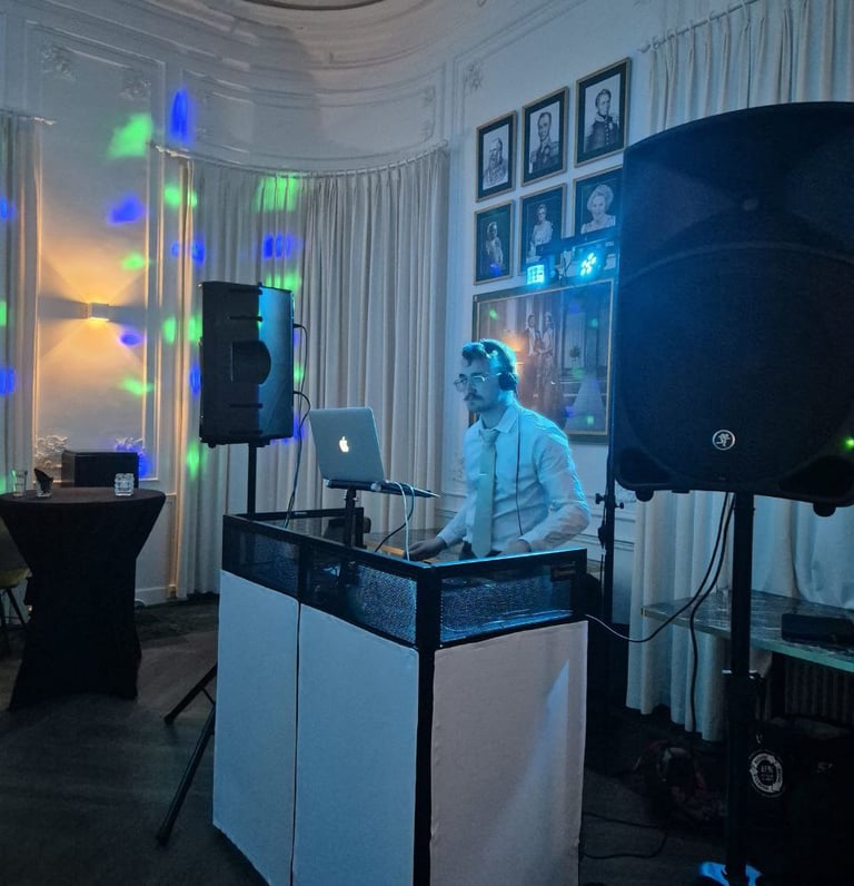 Professional event DJ with laptop and large speakers performing at a party venue with disco lights.