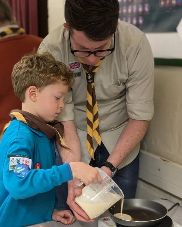 Beaver Scout pouring pancake batter into a pan with the help of a scout leader.