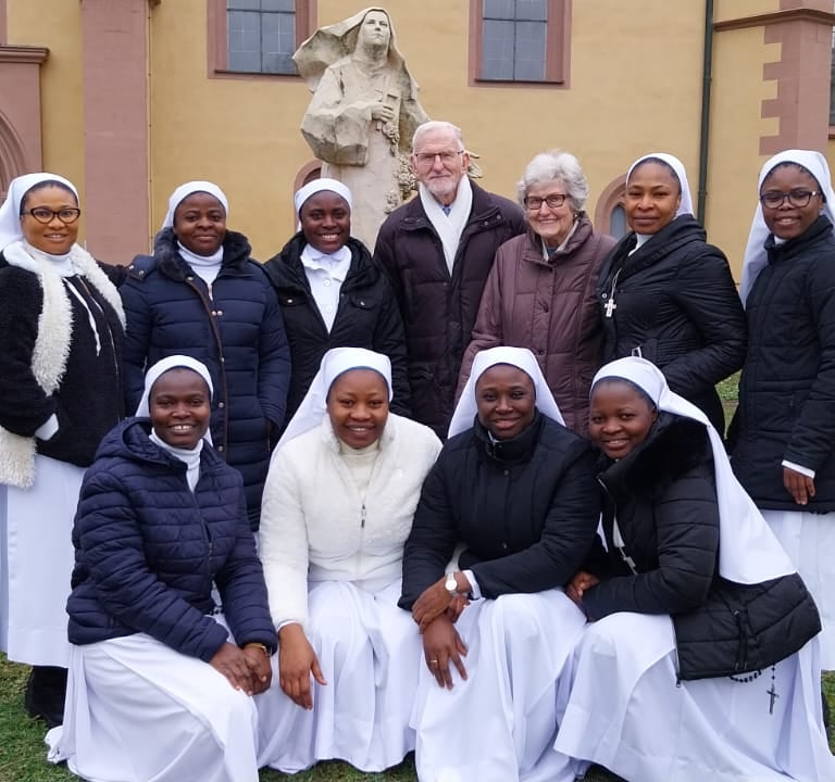 A group of Catholic nuns in white habits and black jackets posing with a senior couple in front of a church.