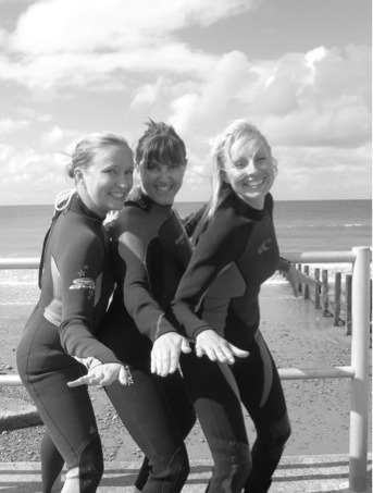 3 women in wetsuits posing in surfer stances next to the beach.