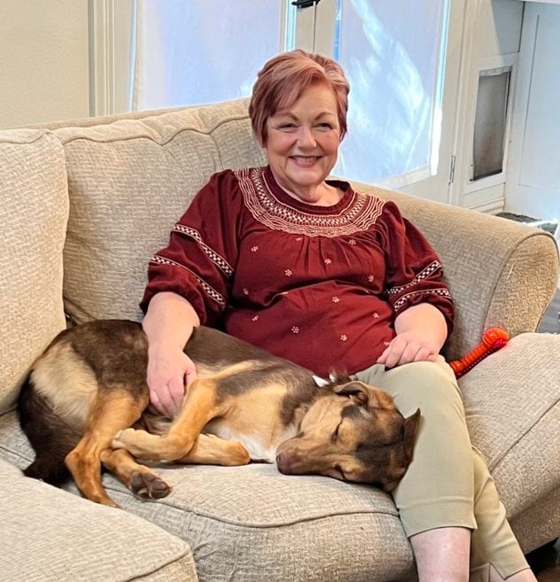 Photo of Debbie, seated on the couch with a resting medium-sized, brown and tan dog.