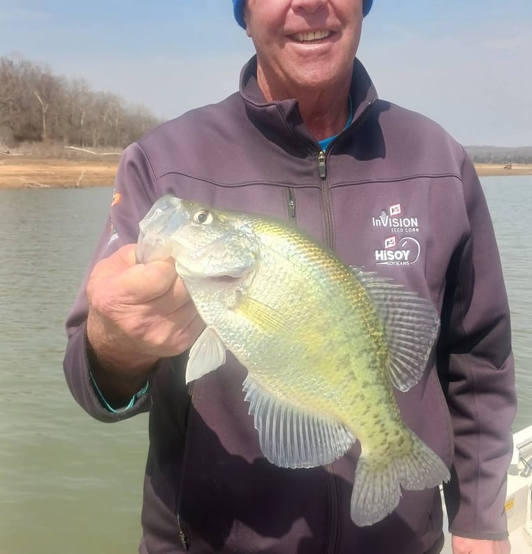 A smiling fisherman on a boat holding a large freshly caught crappie fish over the lake water.