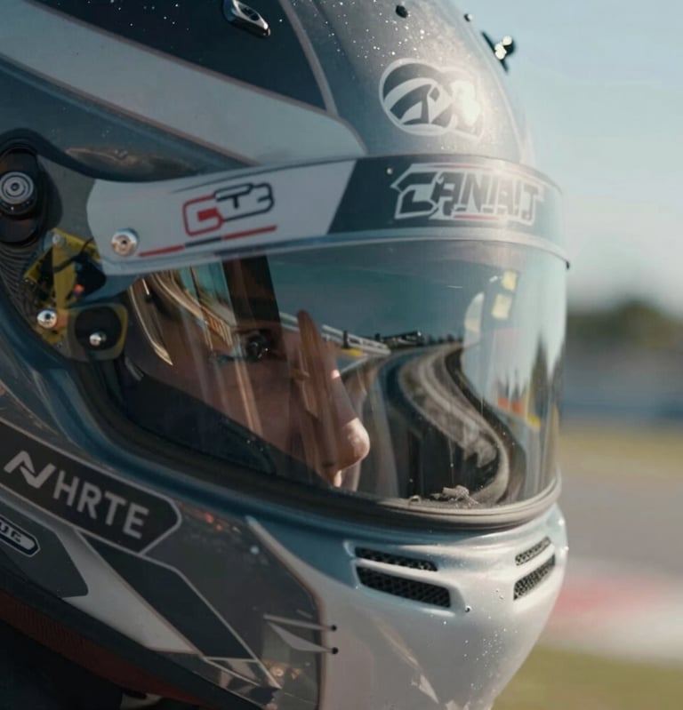 A macro shot focusing on the visor of a racing driver's helmet. The dark gray visor reflects the distorted curve of a famous North American / US racetrack under a clear sky. Intimate and intense focus.