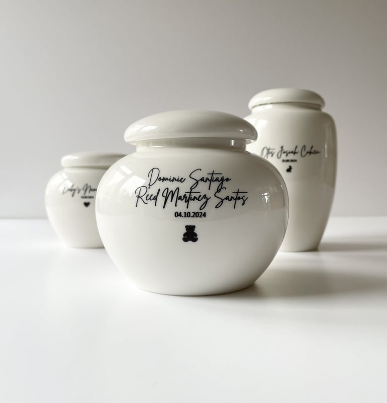 Set of three glossy white ceramic baby urns, each engraved with names, dates, and tiny black heart or bear icons.