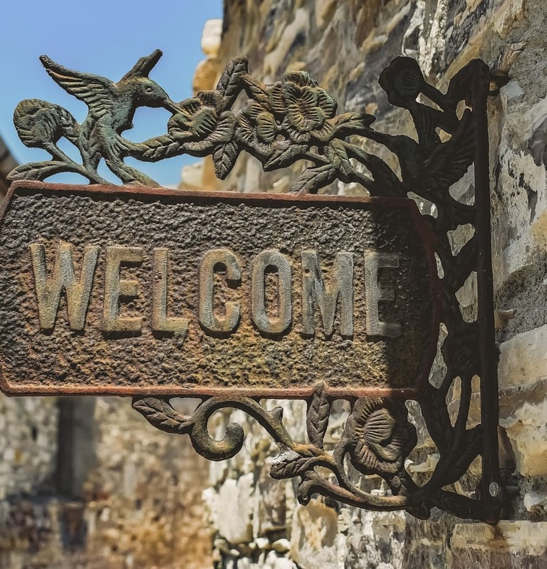 An ornate cast iron "WELCOME" sign, featuring a hummingbird and floral designs, hangs from a stone wall.