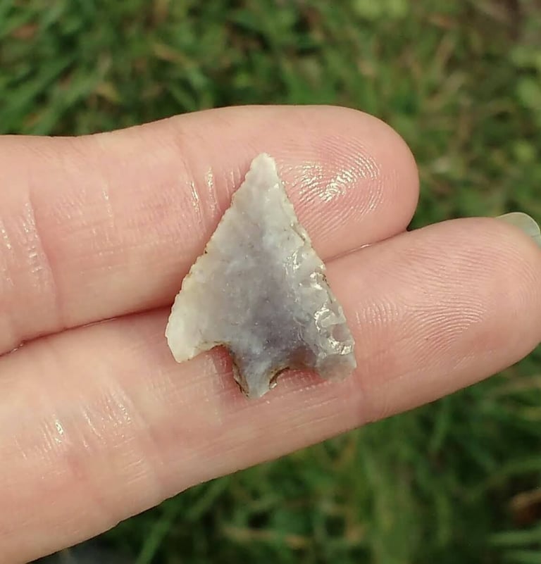 A tiny arrow shaped flint weapon, light grey, held on two fingers.