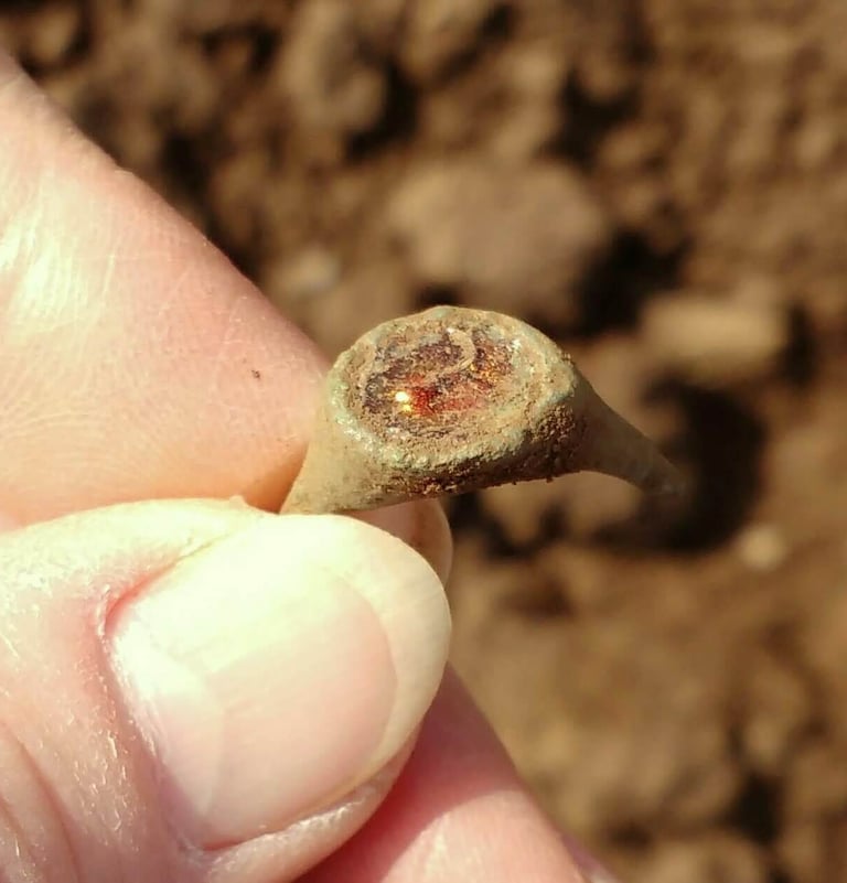 A broken bronze finger ring with green patina and orange inset stone, held between finger and thumb.