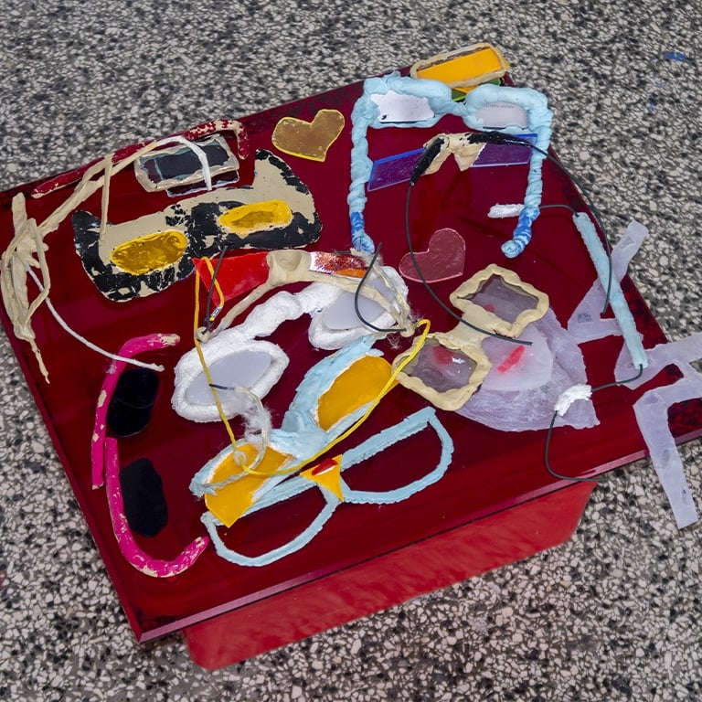 Collection of handmade artistic sunglasses and paper frames on a red glass table surface.