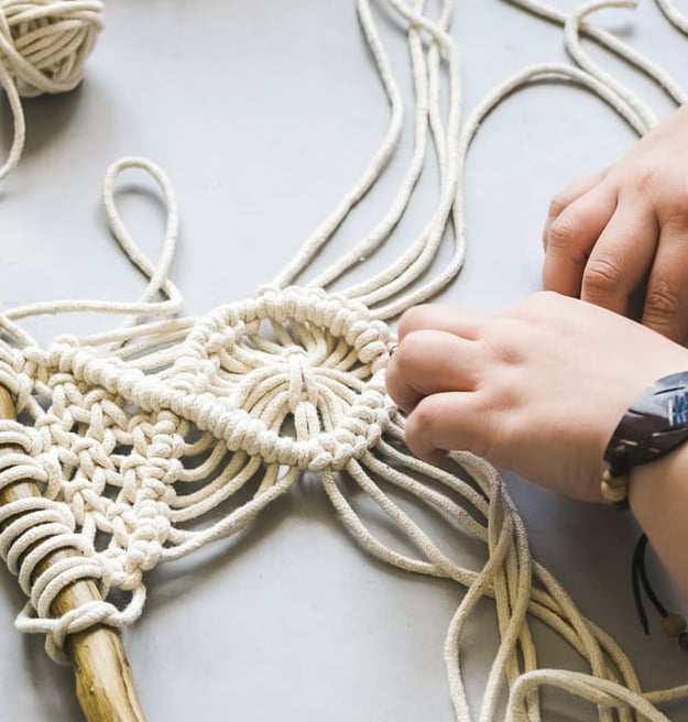 Manos elaborando un colgante de pared de macramé usando cordón de algodón blanco y una madera.