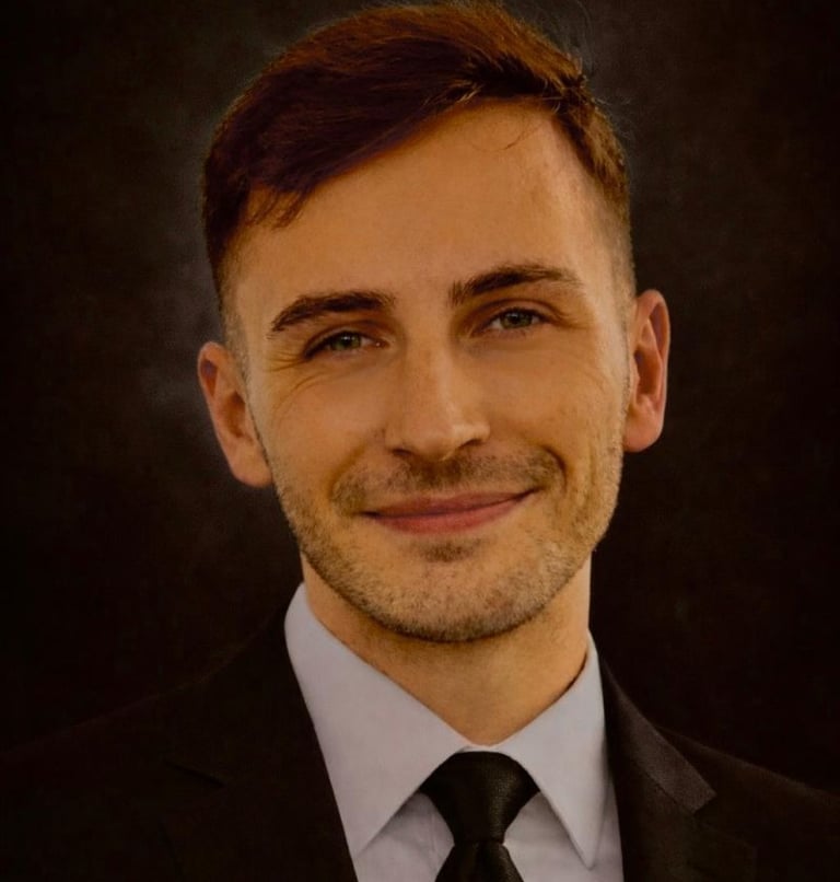 A smiling Bradley in a black suit and tie against a dark background.