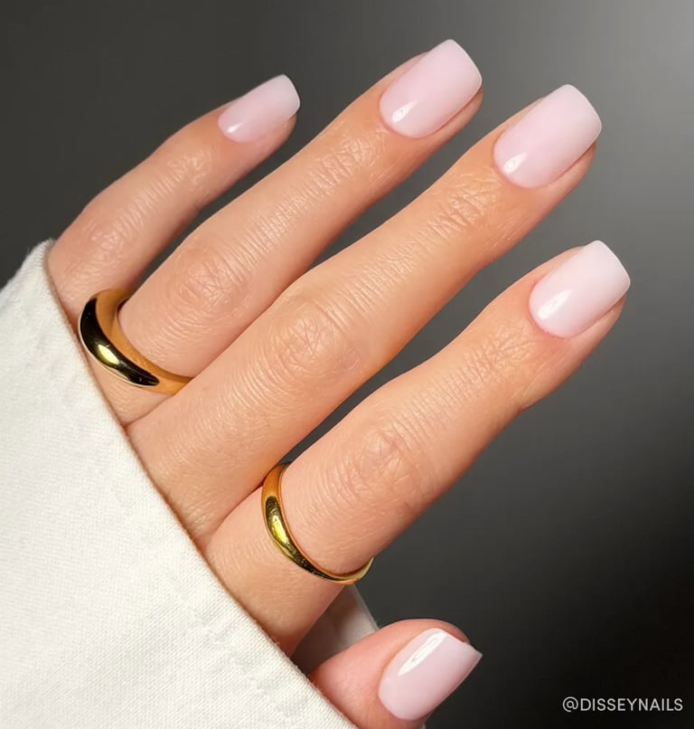 Elegant pale pink manicure on short square nails with gold rings on a person's hand.