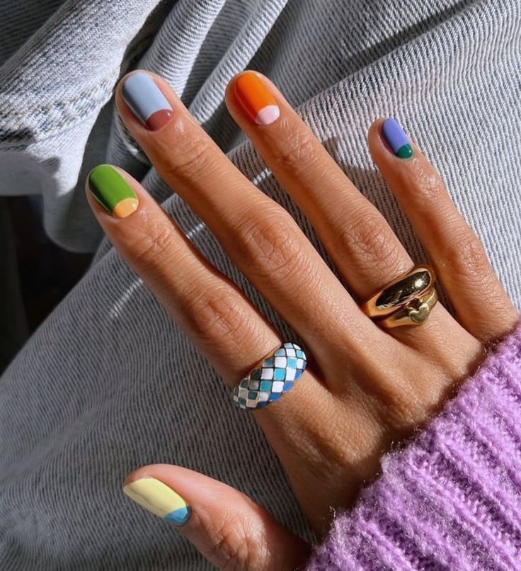 Hand with colorful geometric nail art design and gold rings against denim and a purple sweater.