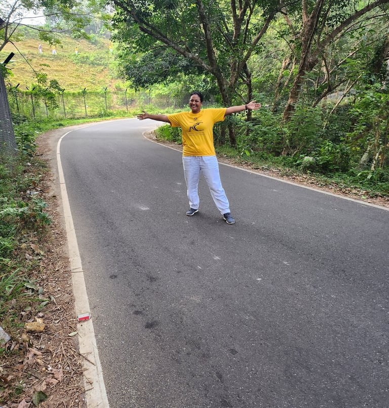 A smiling woman in a yellow shirt stands with arms open on a winding paved road through a lush green forest.